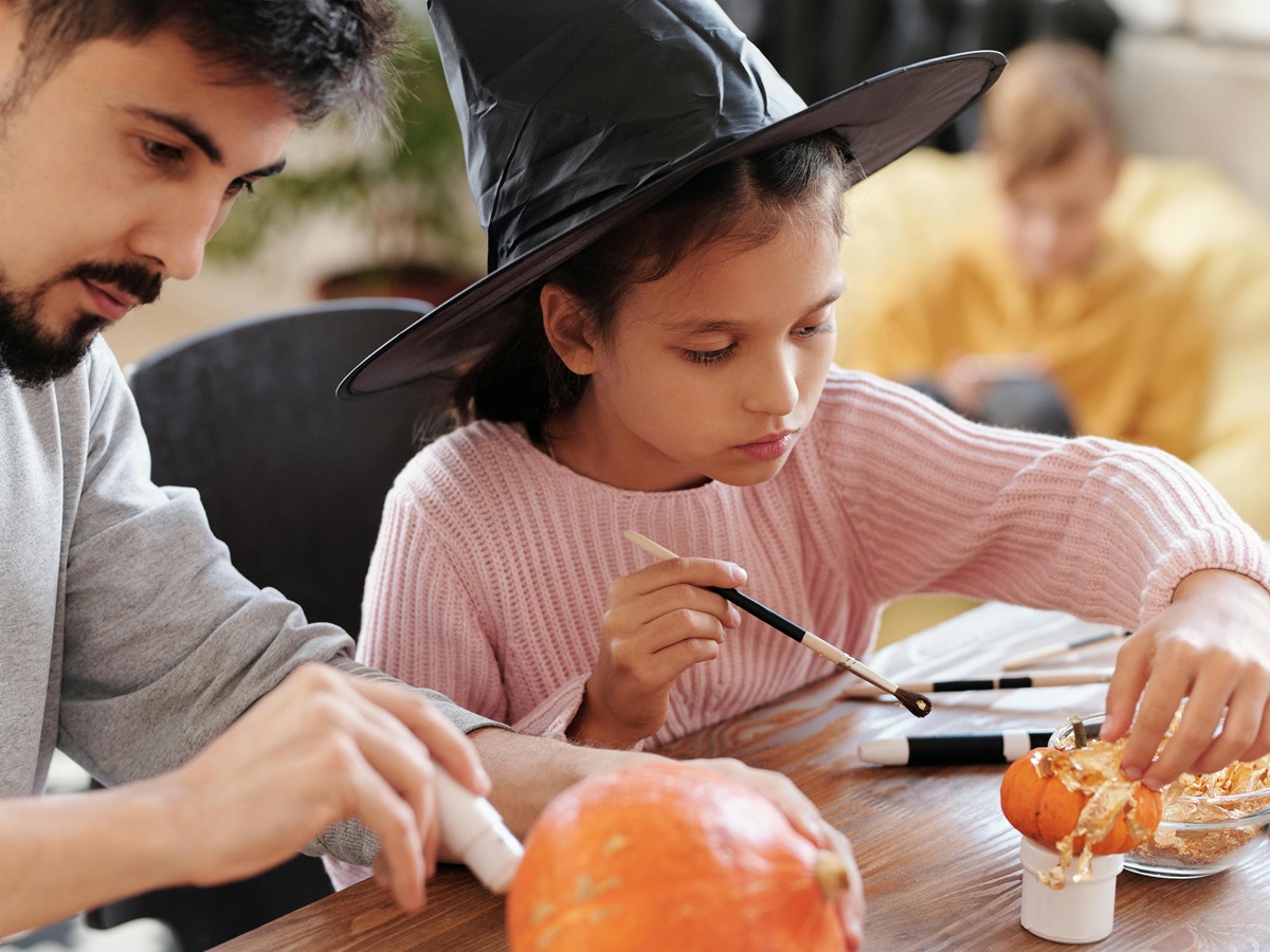 Father and daughter painting Halloween pumpkins together, the little girl wearing a witch hat, illustrating Halloween French vocabulary and crafts.